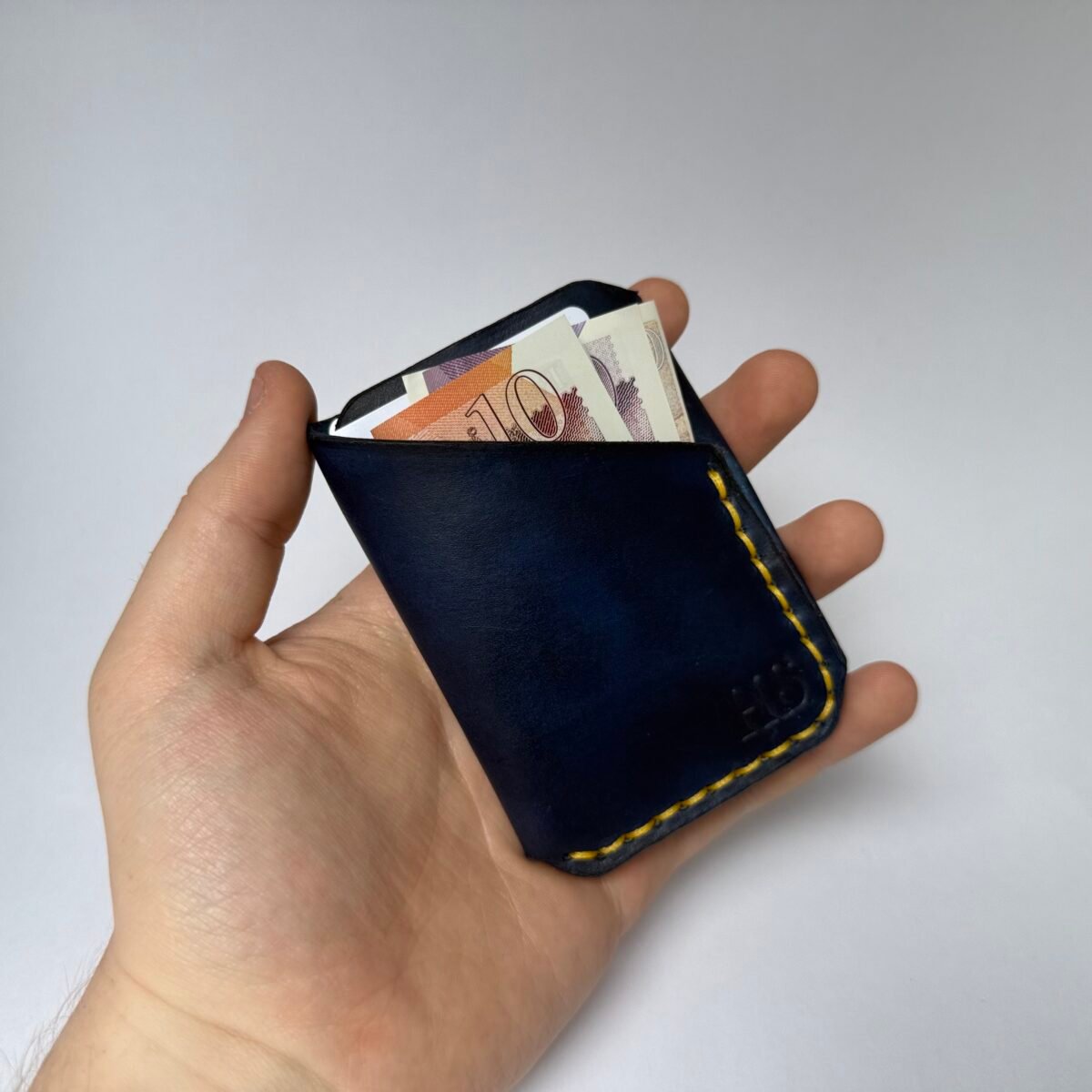Handmade navy blue leather wrap wallet with yellow stitching shown in hand with cash
